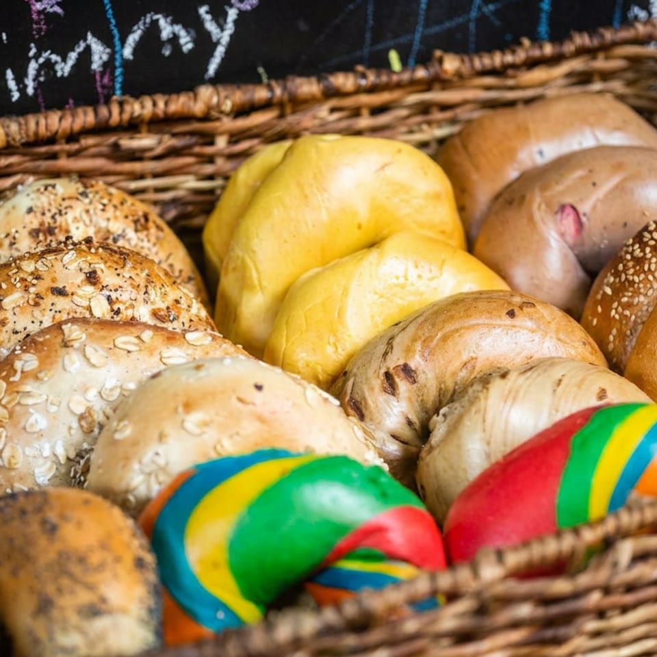 Wicker basket filled with assorted artisan breads, seeded rolls, bagels, and brightly painted multicolored loaves.