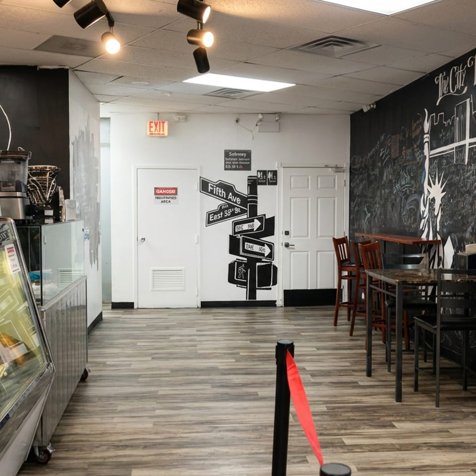 Café interior with pastry display cases, seating, snack shelves, and a chalkboard mural featuring New York landmarks.