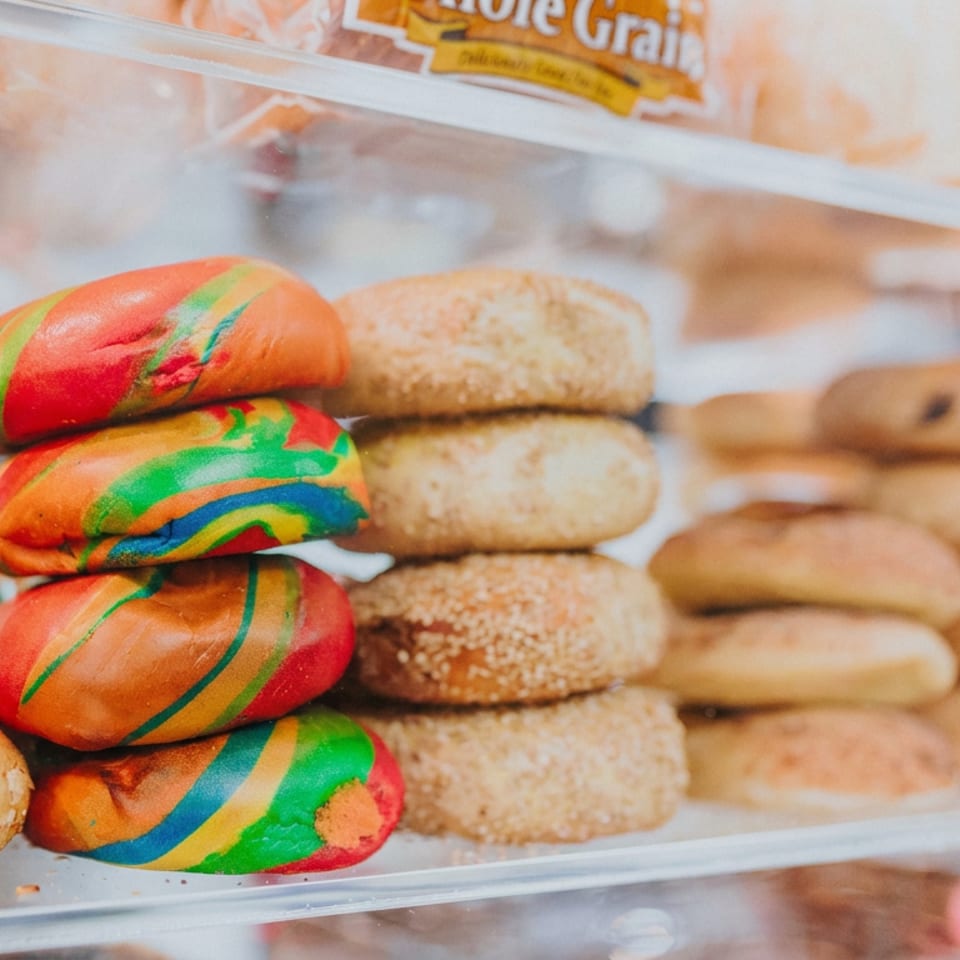 Colorful rainbow bagels stacked beside sesame bagels and assorted plain bagels in a bakery display case.